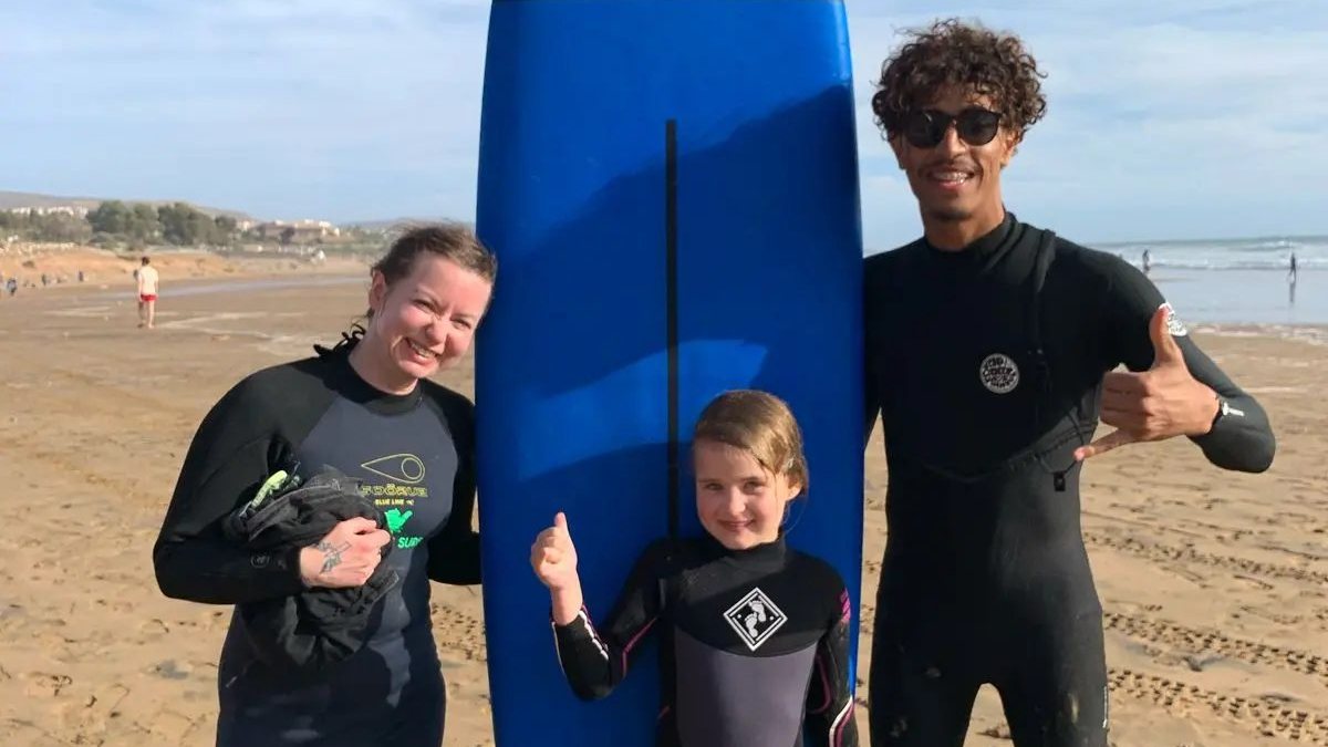 Reda with a smiling young girl and her mother next to a blue soft-top surfboard on the beach after a family surf lesson