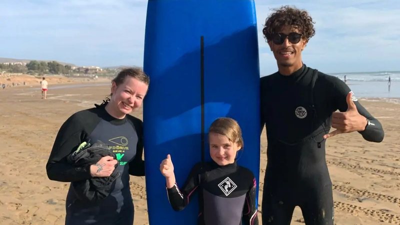 Reda with a smiling young girl and her mother next to a blue soft-top surfboard on the beach after a family surf lesson