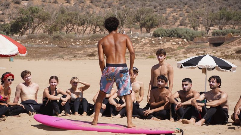Reda standing on a pink soft-top board on the sand, showing surf students how to pop up before they go in the water