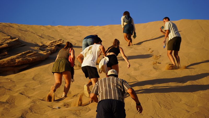 Group climbing the big golden dune at Timlaline near Taghazout under a blue sky