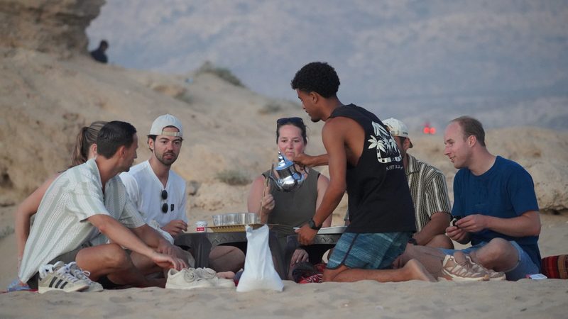 Local guide pouring Moroccan mint tea for the group sitting on rugs on the dunes