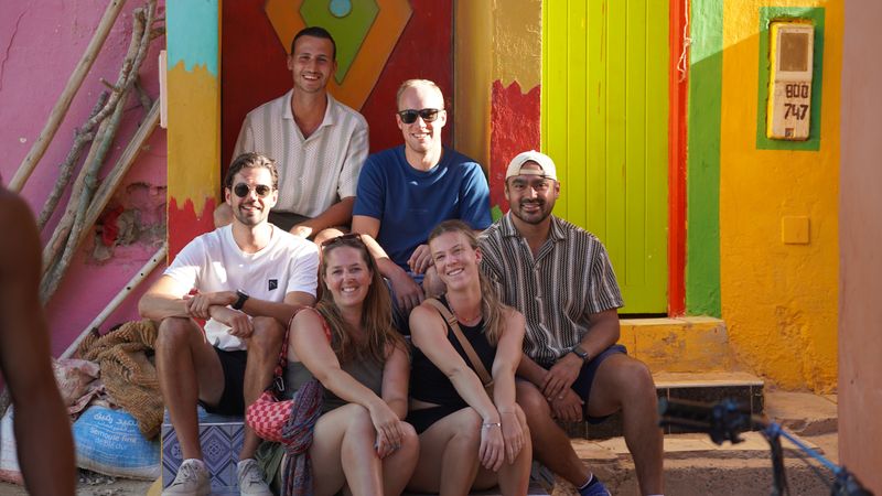 Sandboarding group sitting on the steps of a brightly painted house in a colourful Moroccan village
