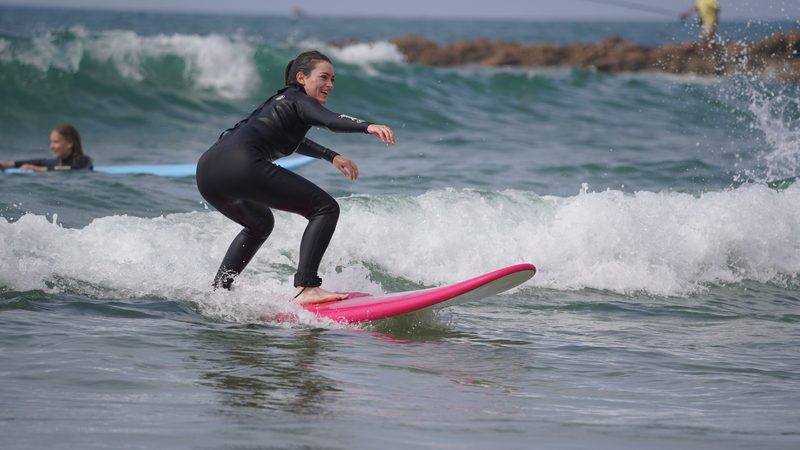 Smiling surf student riding a whitewater wave on a pink soft-top board during a beginner lesson near Taghazout
